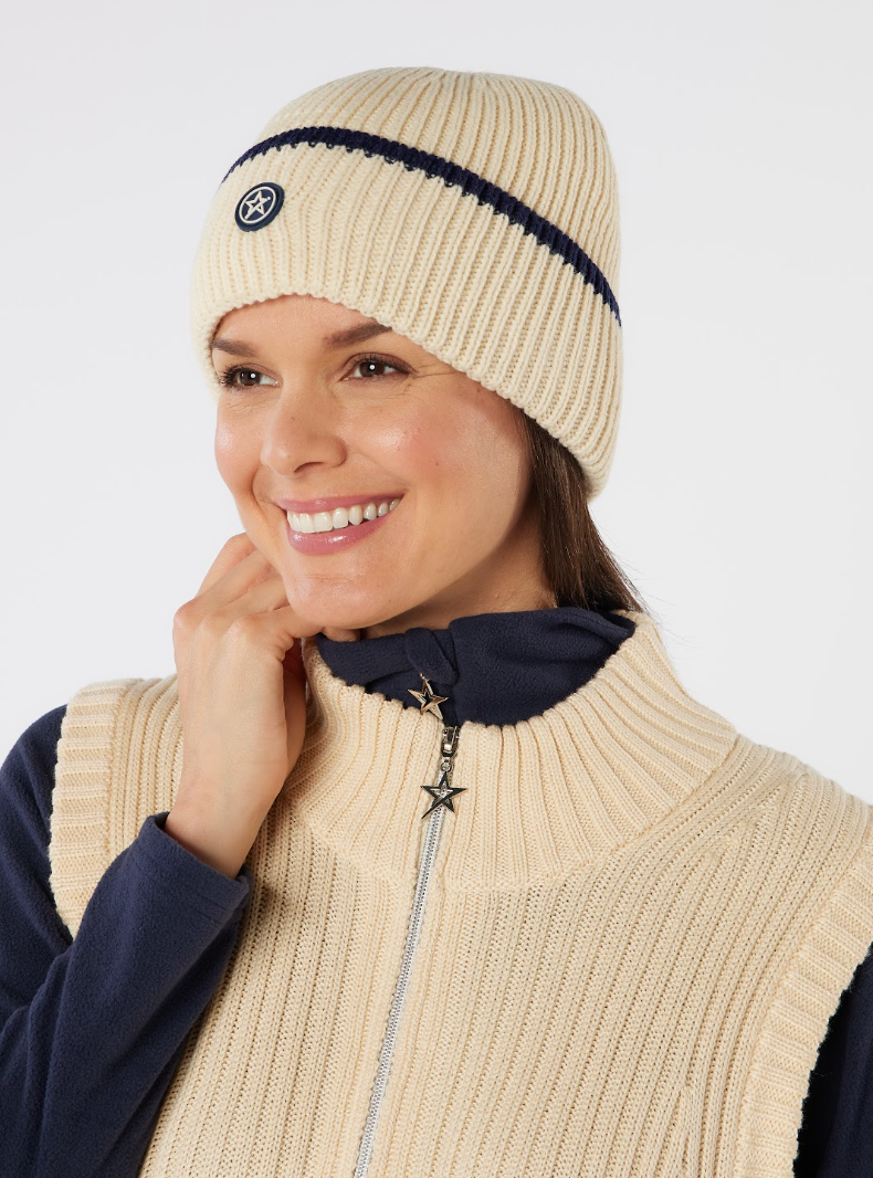 A smiling woman wearing a cream ribbed knit golf beanie with a navy stripe and logo, paired with a matching cream zip-front vest over a navy top.