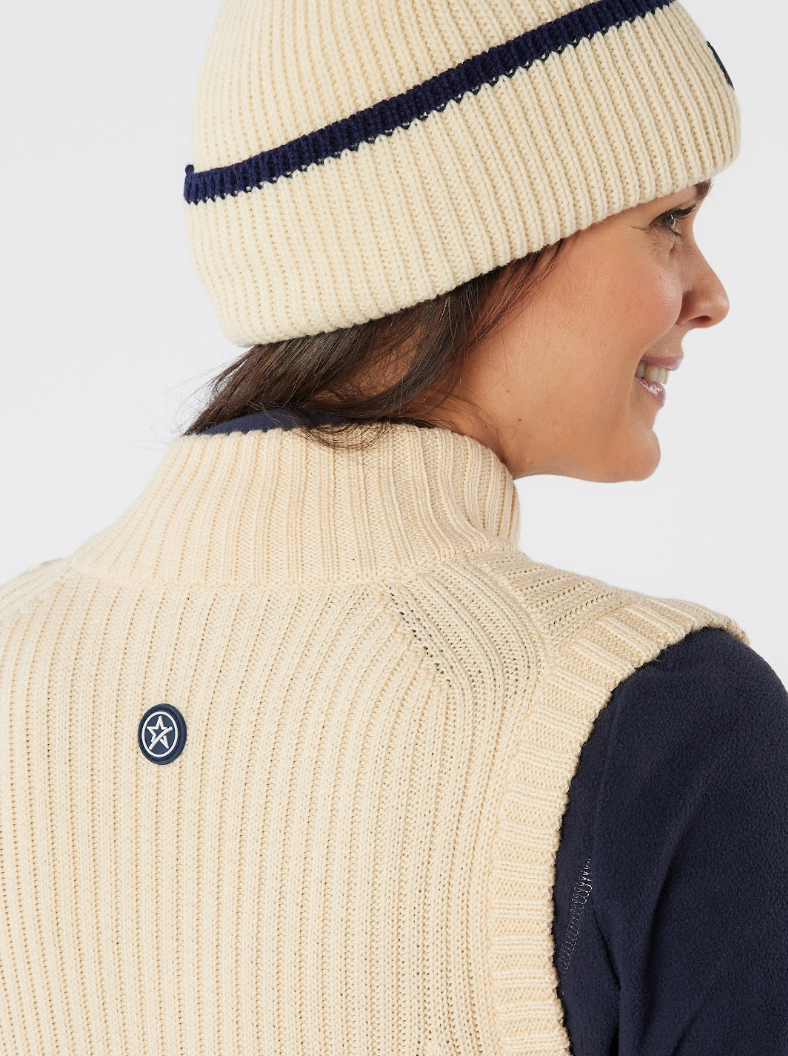 A smiling woman wearing a cream ribbed knit golf beanie with a navy stripe, paired with a matching cream vest.
